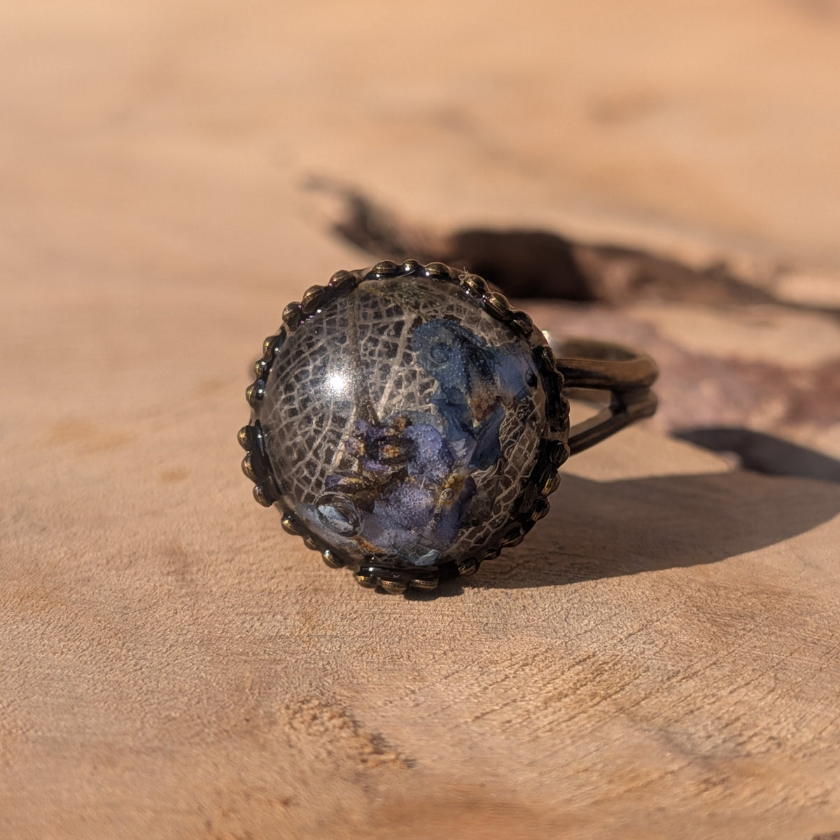 Blue forget-me-not and white skeleton leaf resin ring resting on sunlit wood, framed by a decorative rustic brown border, EL-Creations