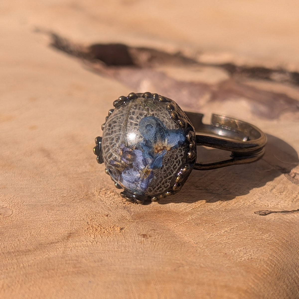 Angled view of a blue forget-me-not resin ring on wood, showing white skeleton leaf veins and its open adjustable brown alloy band, EL-Creations