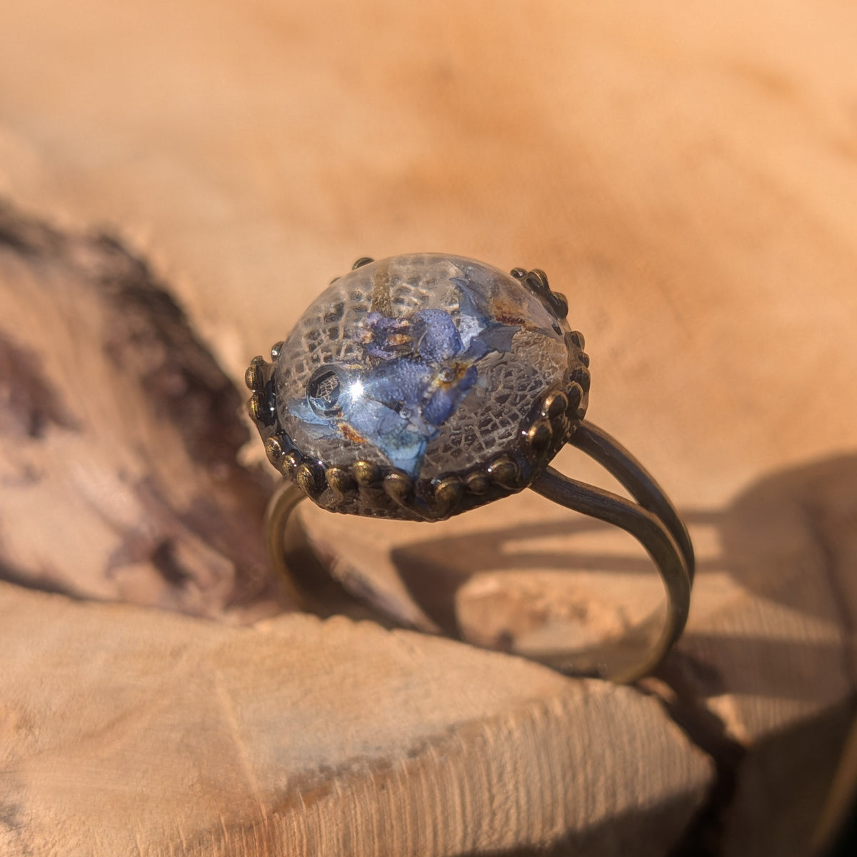 Angled side view of a forget-me-not resin ring with white skeleton leaf, showing its domed cabochon and rustic brown split band on wood, EL-Creations