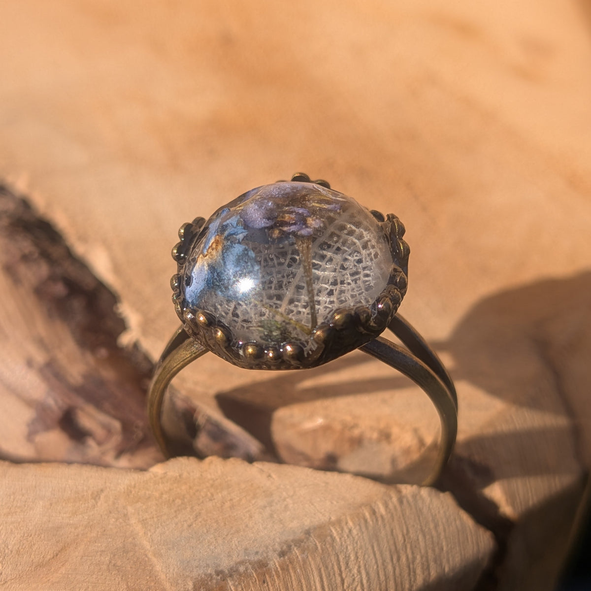 Front view of an adjustable forget-me-not resin ring balanced in a crack in wood, showing its white leaf background and decorative brown setting, EL-Creations