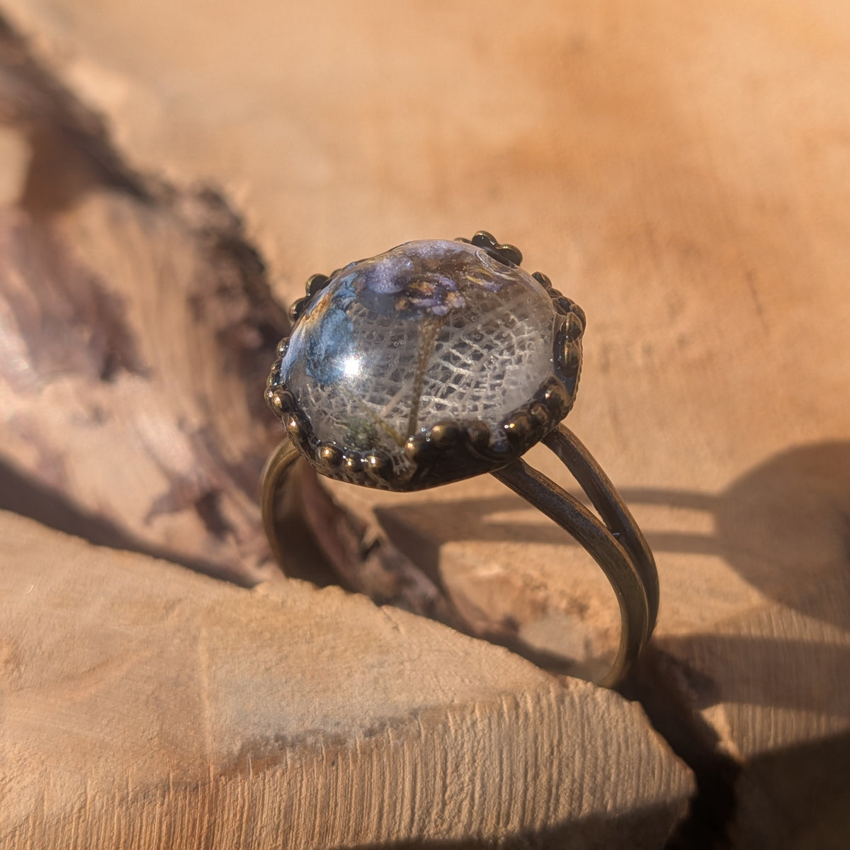 Close-up of a botanical resin ring standing upright on wood, with fine white skeleton leaf veins and blue forget-me-not flowers beneath the glossy dome, EL-Creations