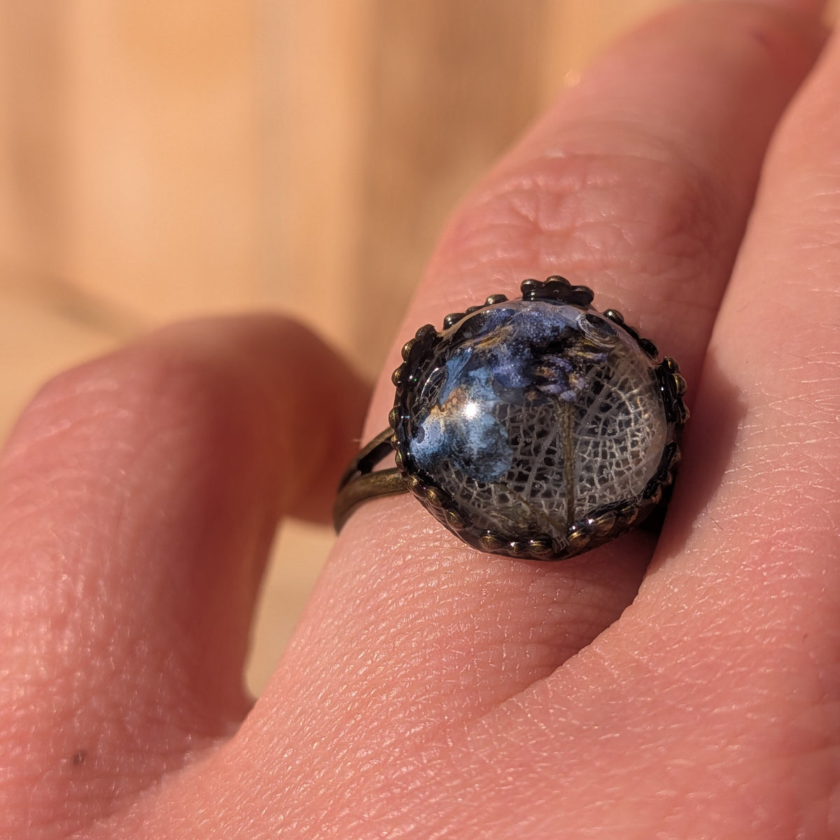 Macro close-up of blue forget-me-not flowers and white skeleton leaf preserved in a resin ring worn on a finger in sunlight, EL-Creations
