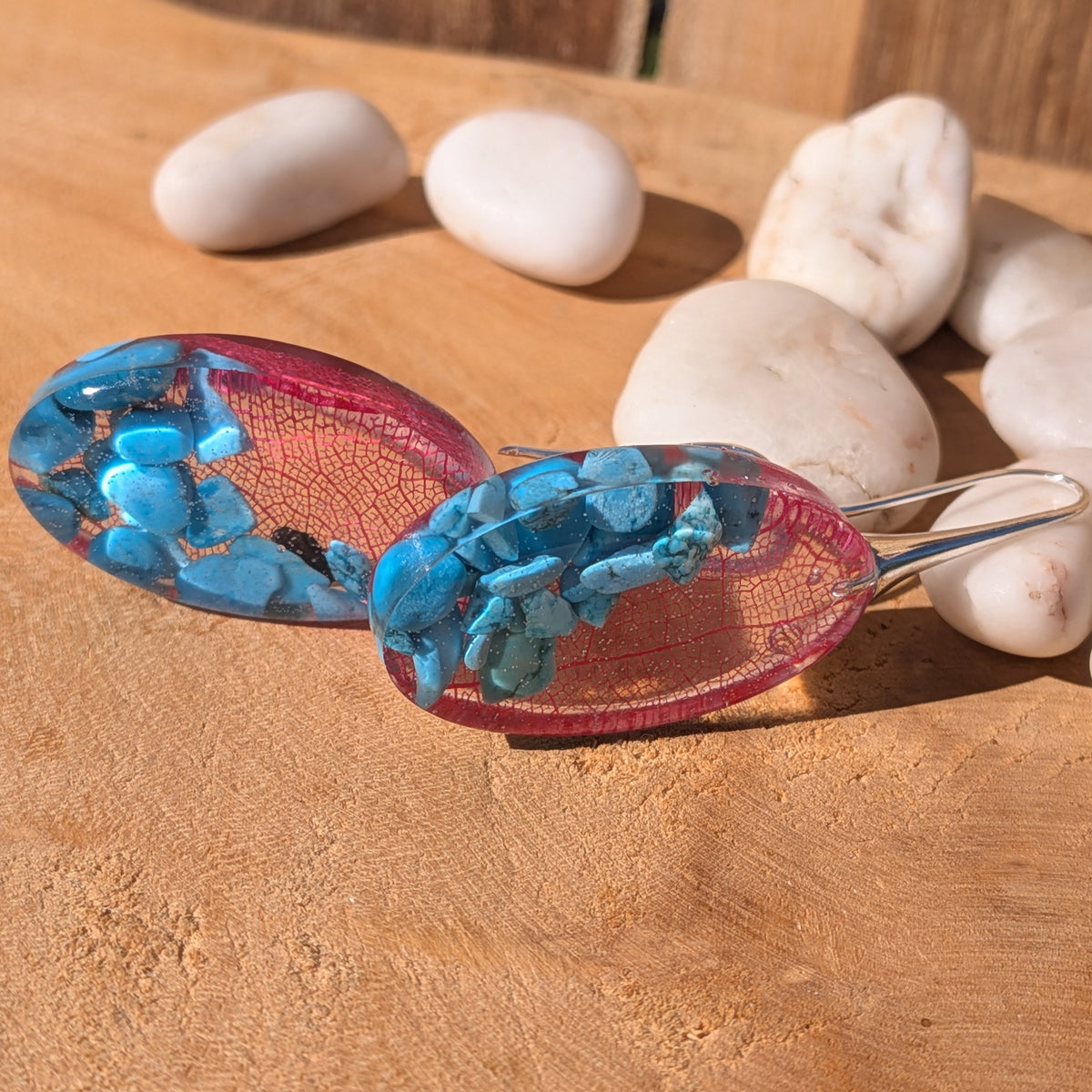 Low-angle view of oval resin earrings showing embedded turquoise chips and red skeleton leaves, EL-Creations