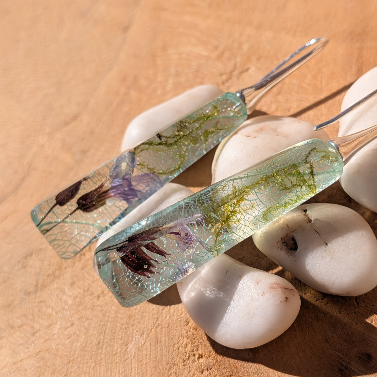 Slender resin earrings with light blue skeleton leaves and green moss beside white pebbles, EL-Creations