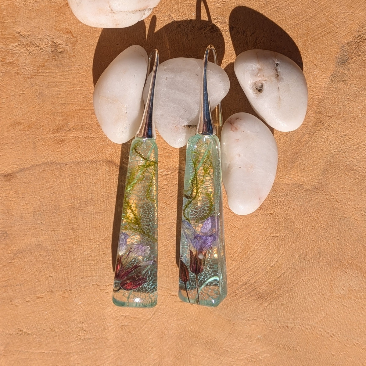 Overhead view of resin drop earrings with moss and light blue skeleton leaves beside white pebbles, EL-Creations