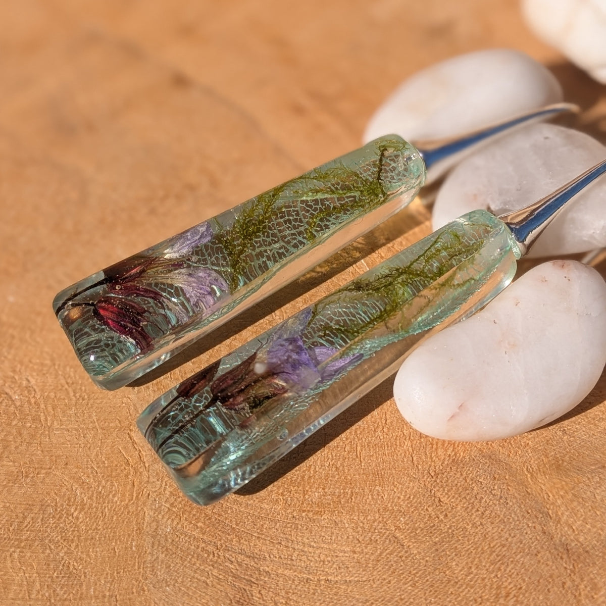 Pair of elongated botanical resin earrings lying on wood with silver-tone hooks, EL-Creations