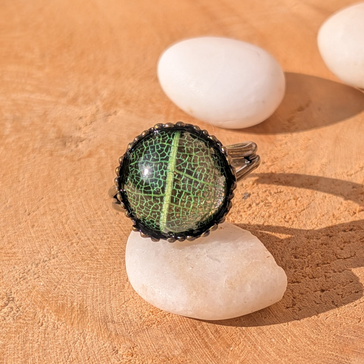 Forest Lace resin ring with fine green leaf veins in a rustic brown setting resting on a white pebble on sunlit wood, EL-Creations