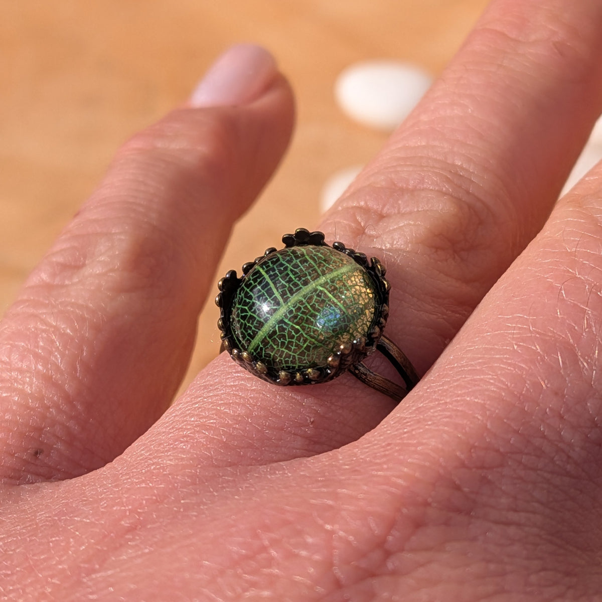 Angled close-up of a green leaf vein resin ring worn on a finger in sunlight, with its decorative rustic brown border visible, EL-Creations