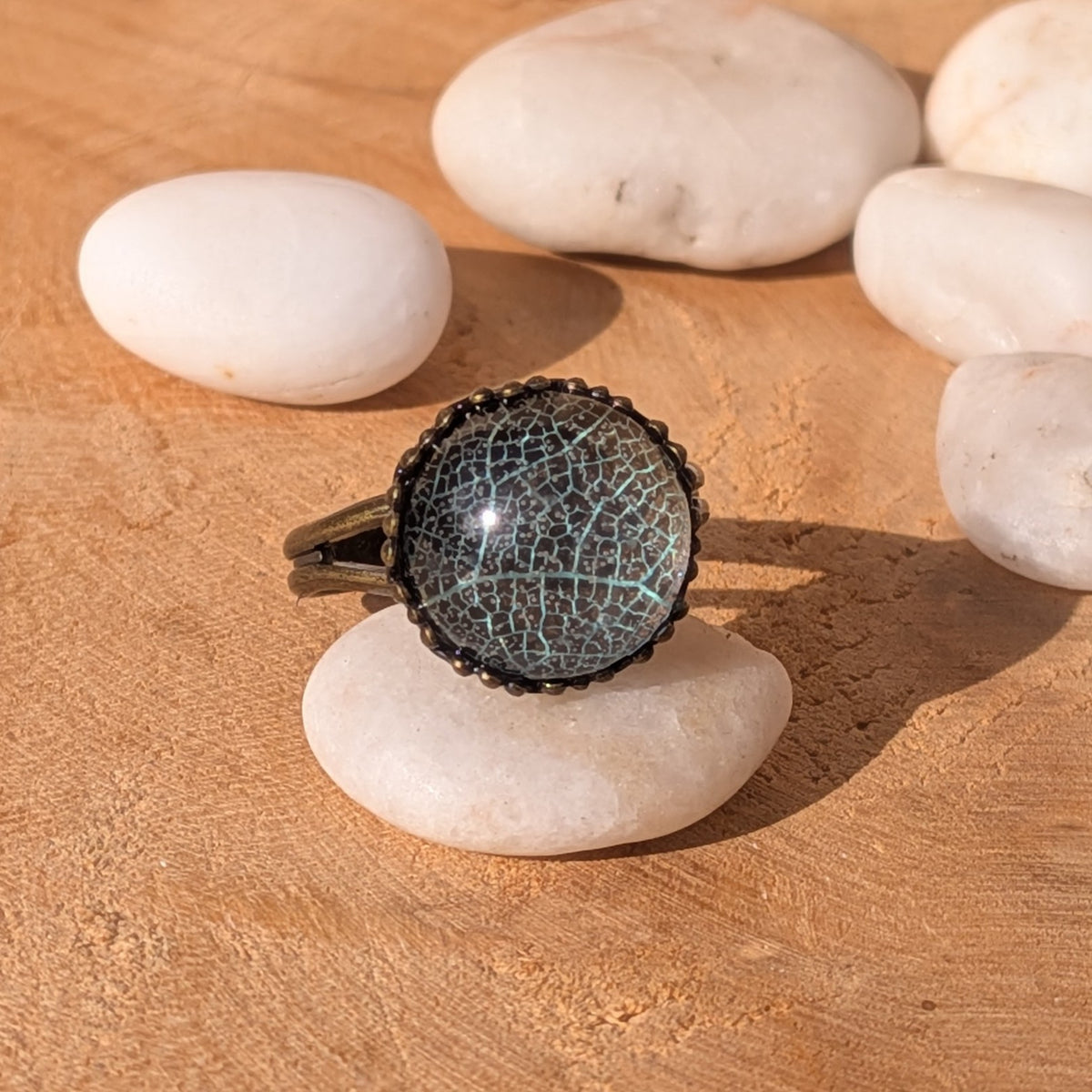 Pale turquoise-green leaf resin ring displayed on a white stone among pebbles, with its decorative brown border visible in sunlight, EL-Creations