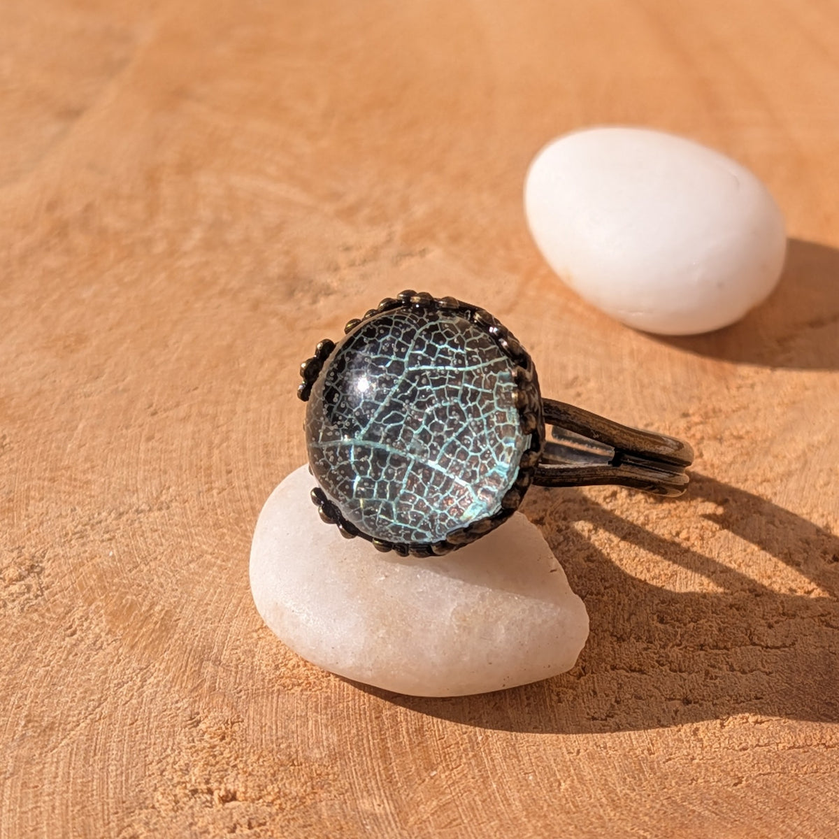 Seafoam Lace resin ring with pale turquoise-green leaf veins in a rustic brown setting resting on a white pebble on sunlit wood, EL-Creations