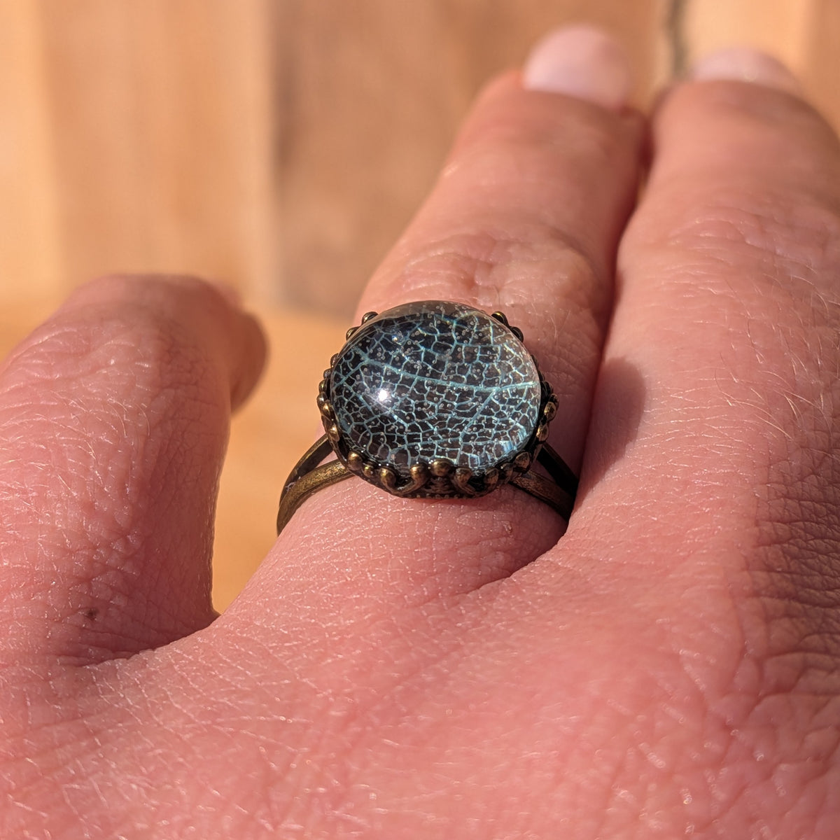 Close-up of an adjustable Seafoam Lace resin ring worn on a finger, showing its glossy cabochon and decorative rustic brown setting, EL-Creations