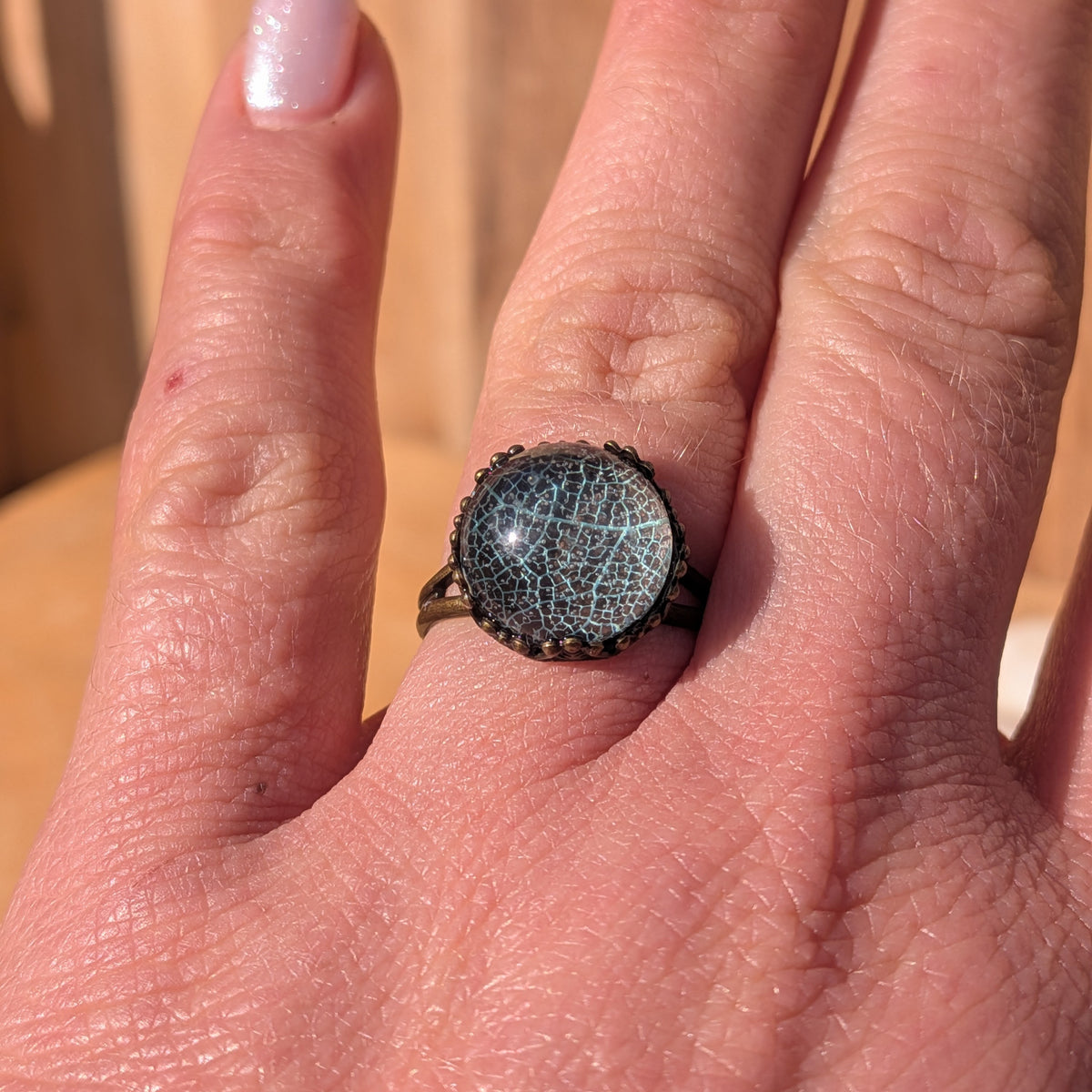 Front view of a pale turquoise-green botanical resin ring worn on an outstretched hand against a wooden background, EL-Creations