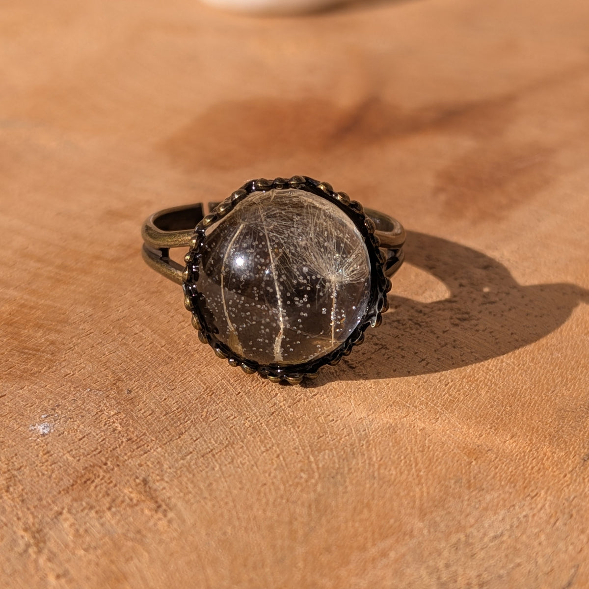 Front view of a handmade dandelion resin ring on a wooden surface, with feathery seed fibres visible beneath the glossy dome, EL-Creations