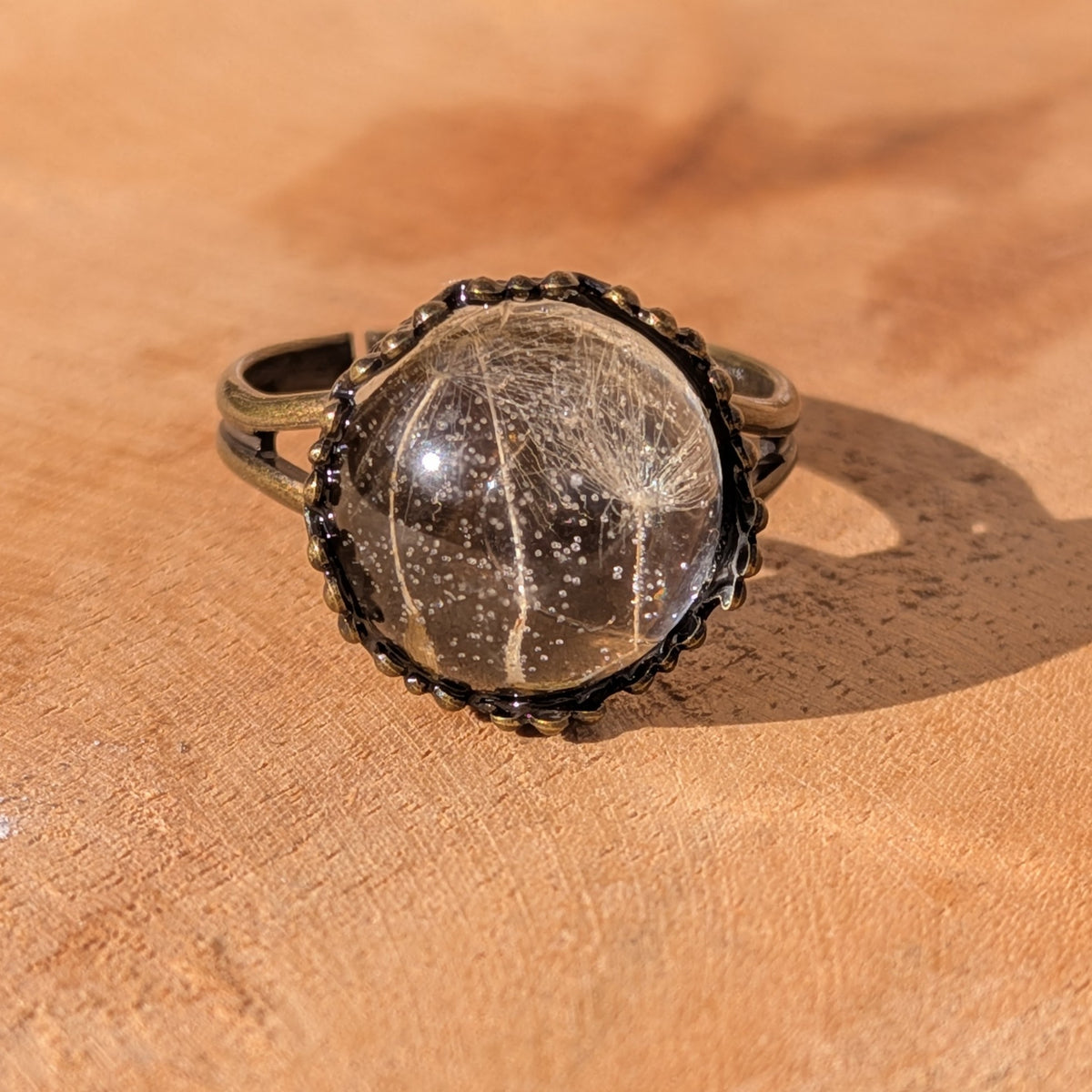 Floating Wishes resin ring with delicate dandelion seeds inside a clear dome, framed by a rustic brown setting on sunlit wood, EL-Creations