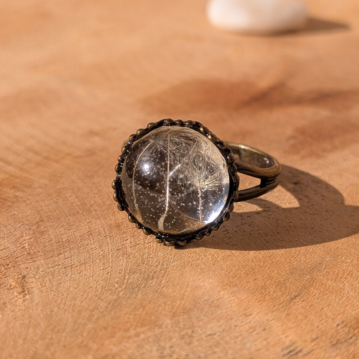 Angled view of a clear dandelion seed resin ring resting on wood, showing its adjustable brown split band, EL-Creations