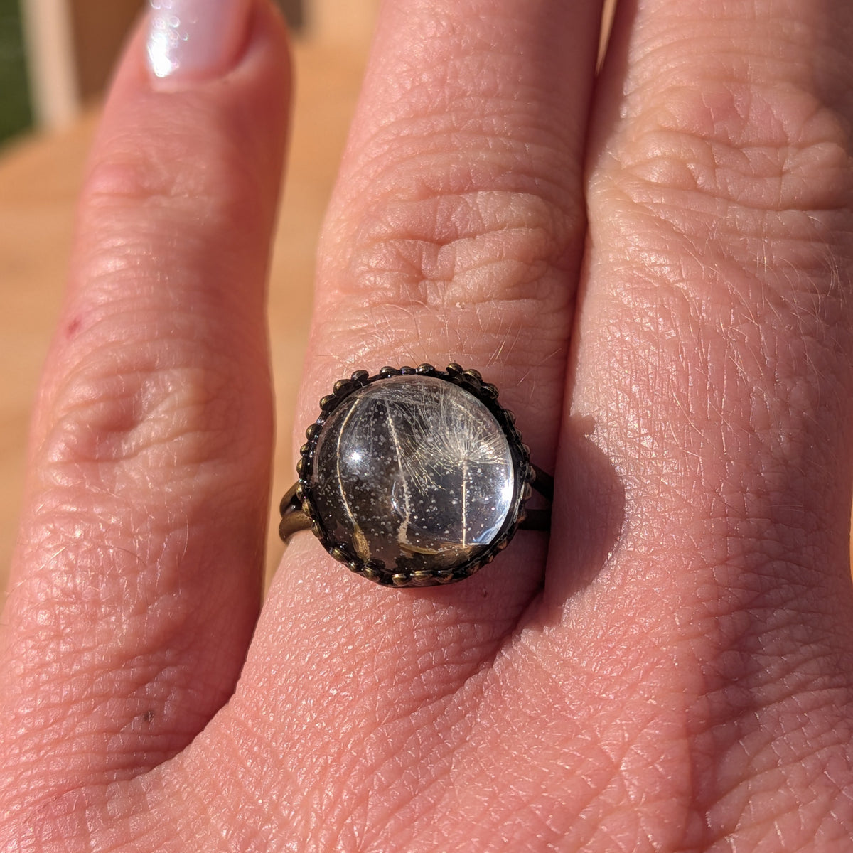 Front close-up of a Floating Wishes dandelion seed resin ring worn on a finger in sunlight, EL-Creations