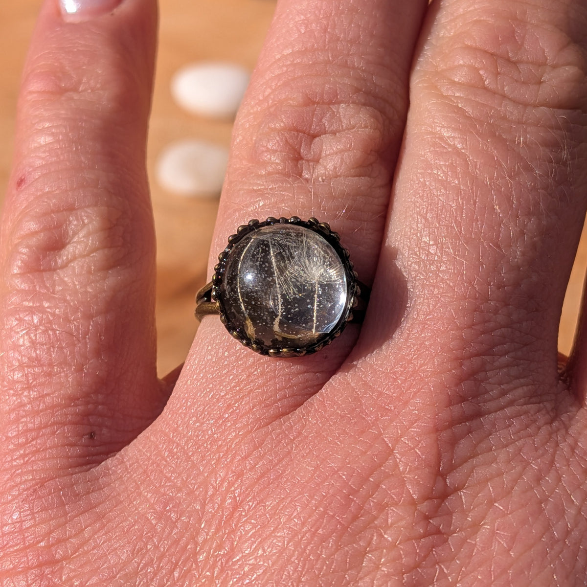 Clear dandelion seed resin ring worn on an outstretched hand, showing its botanical details and rustic brown setting, EL-Creations