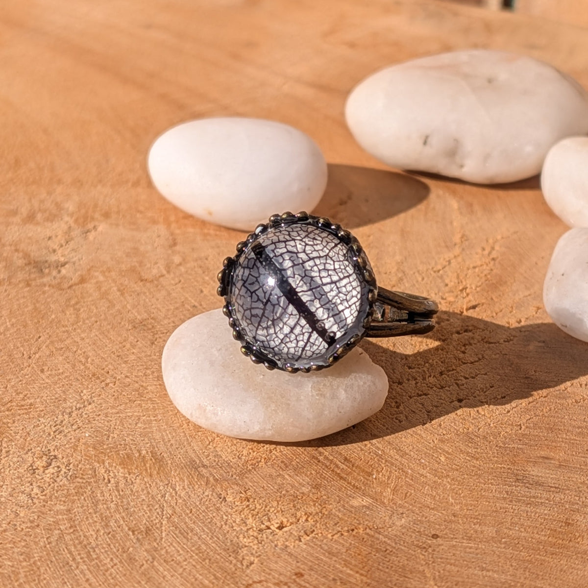 Black skeleton leaf resin ring resting on a white stone among scattered pebbles on sunlit wood, EL-Creations