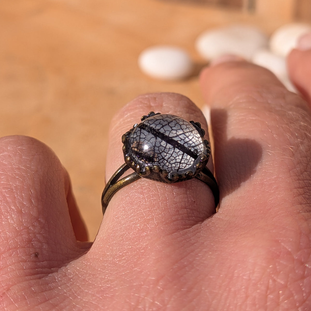 Low side view of a black skeleton leaf ring worn on a finger, highlighting the raised resin dome and decorative metal border, EL-Creations