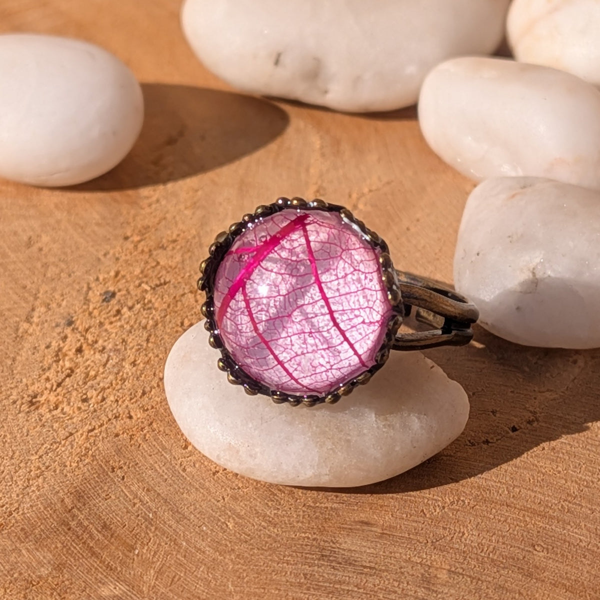 Close-up of a vivid pink skeleton leaf resin ring resting on a white stone, with its rustic brown band visible to the right, EL-Creations