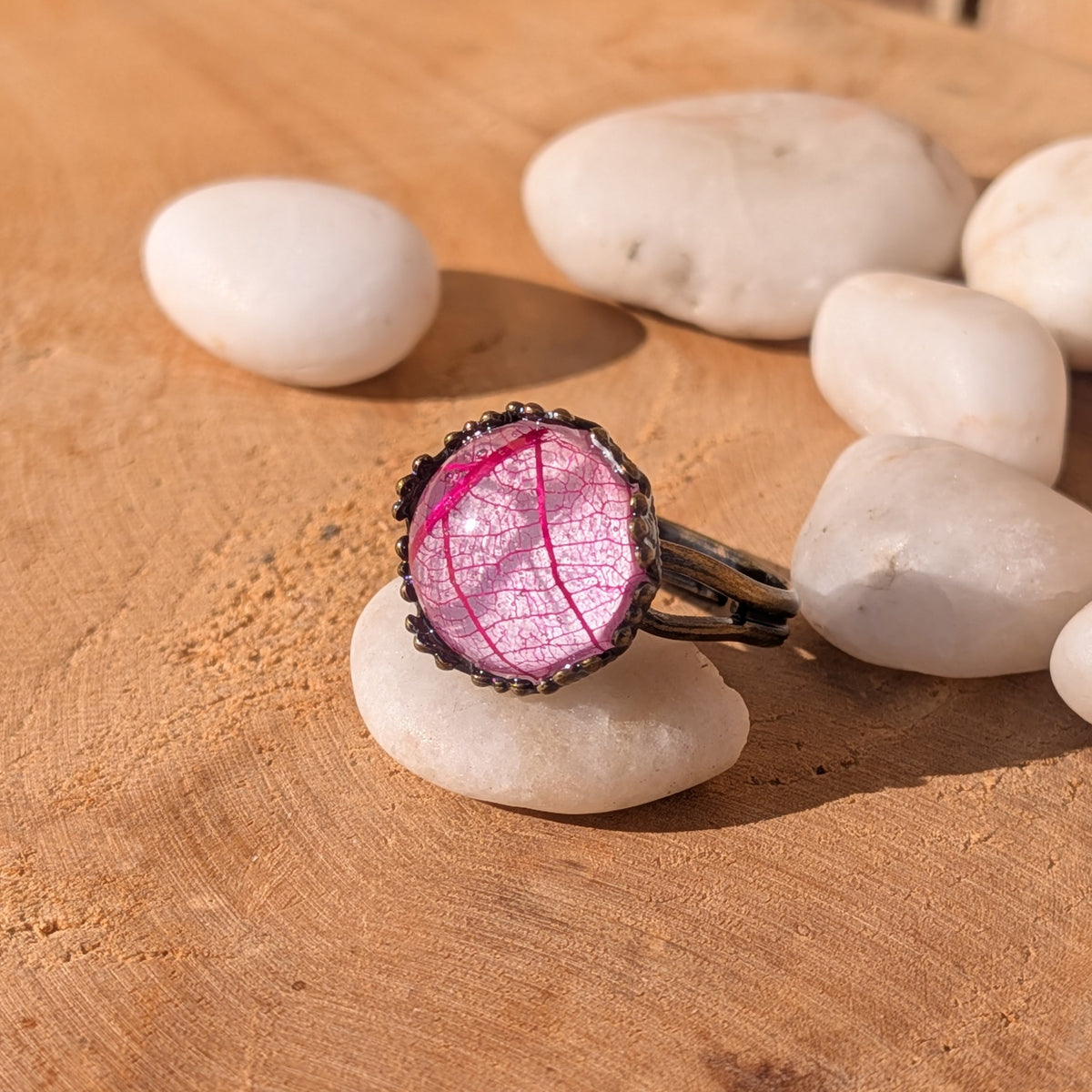 Angled view of a handmade pink botanical resin ring on a white pebble, showing its glossy centre and brown split band, EL-Creations
