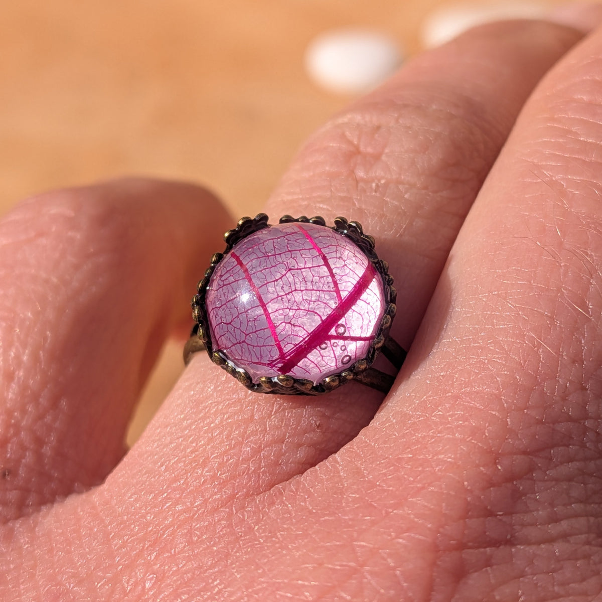 Close-up of a Fuchsia Lace resin ring worn on a finger, showing vivid pink skeleton leaf veins beneath a glossy dome, EL-Creations
