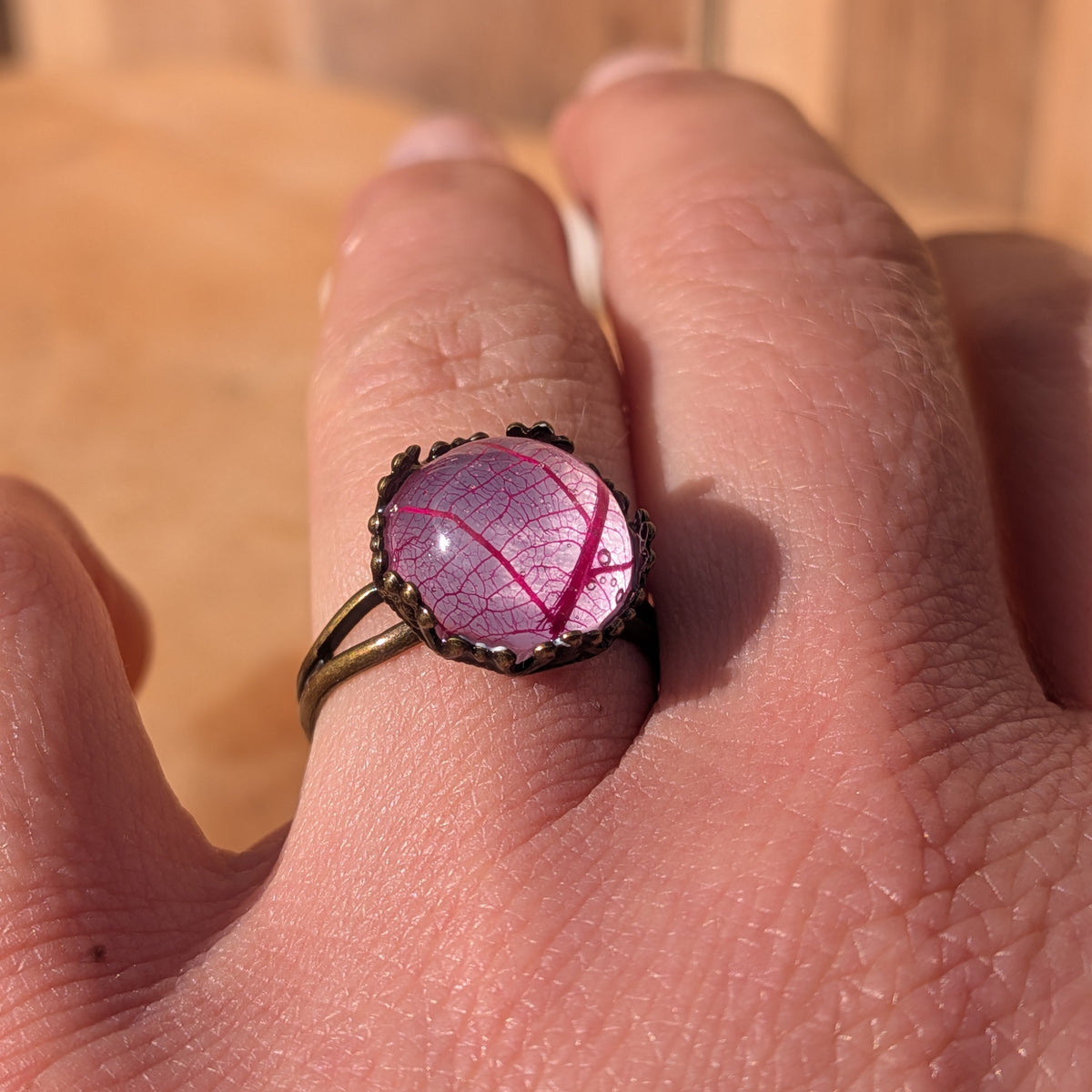 Angled view of a pink leaf resin ring worn on a finger in sunlight, with a decorative rustic brown setting and split band, EL-Creations