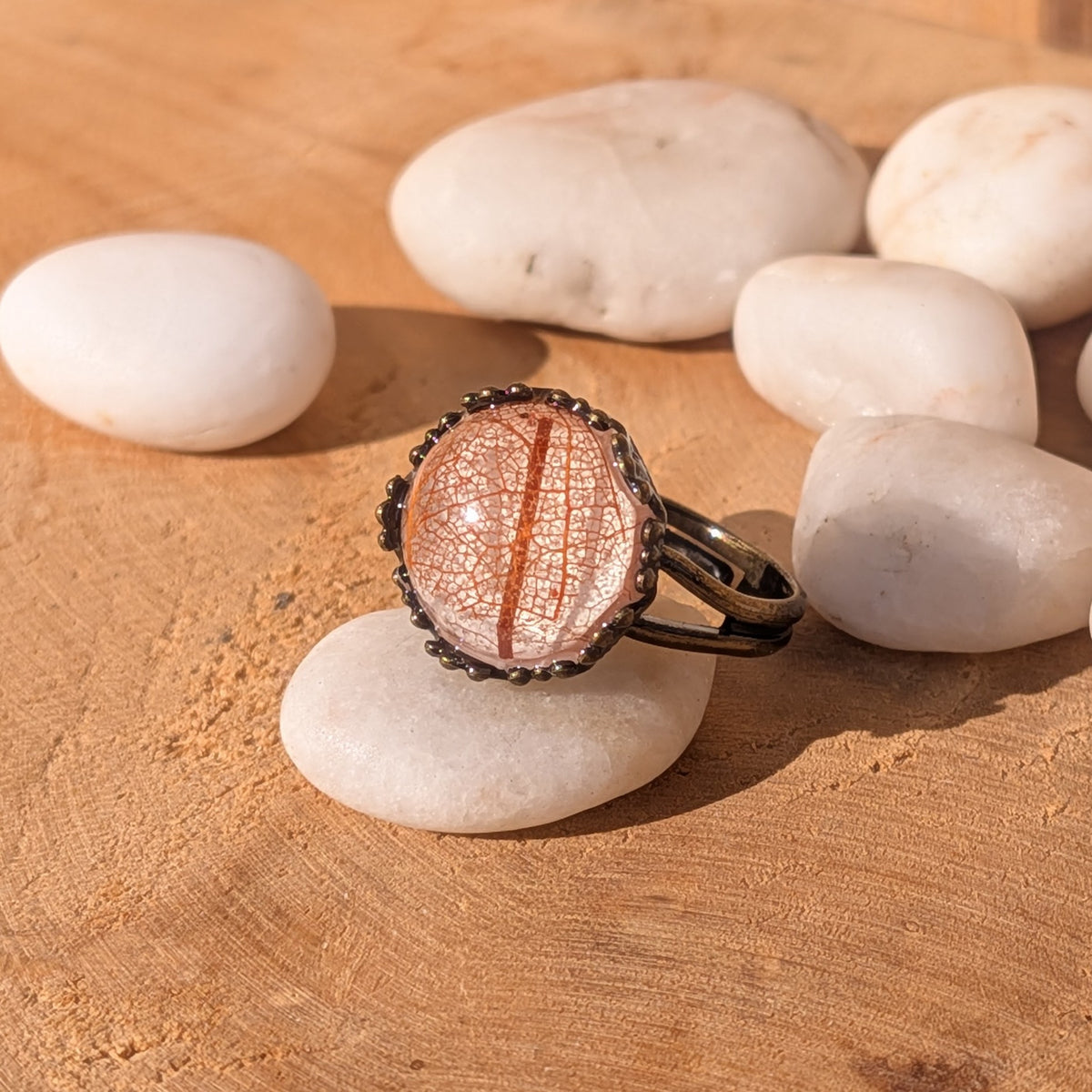 Autumn Glow skeleton leaf resin ring balanced on a white pebble among smooth stones on sunlit wood, EL-Creations