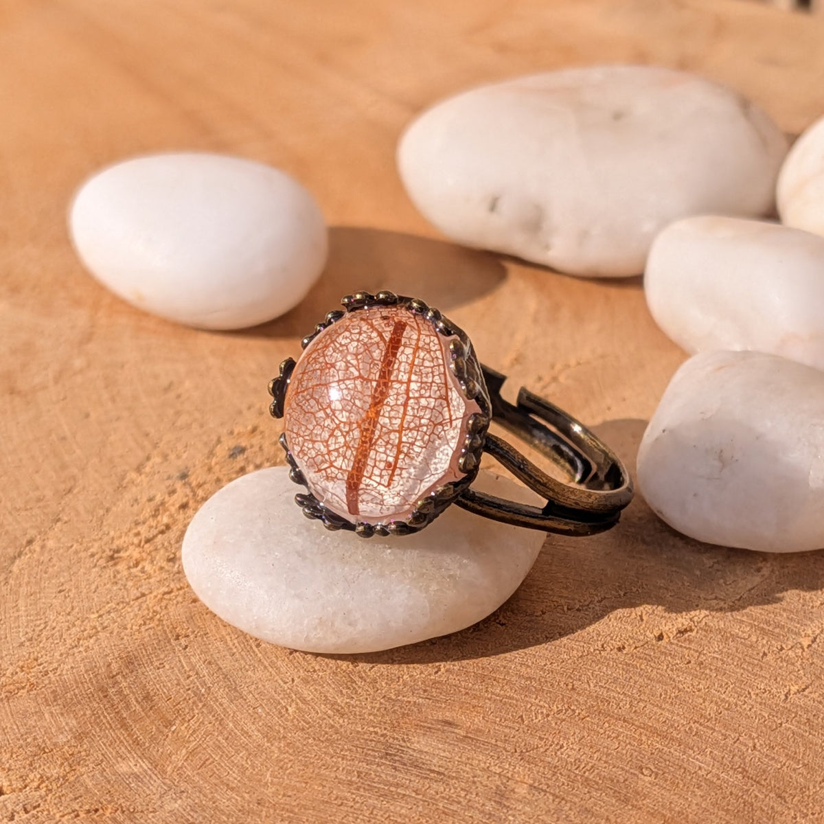 Autumn Glow resin ring with orange and beige skeleton leaf veins, resting on a white pebble with its rustic brown band visible to the right, EL-Creations