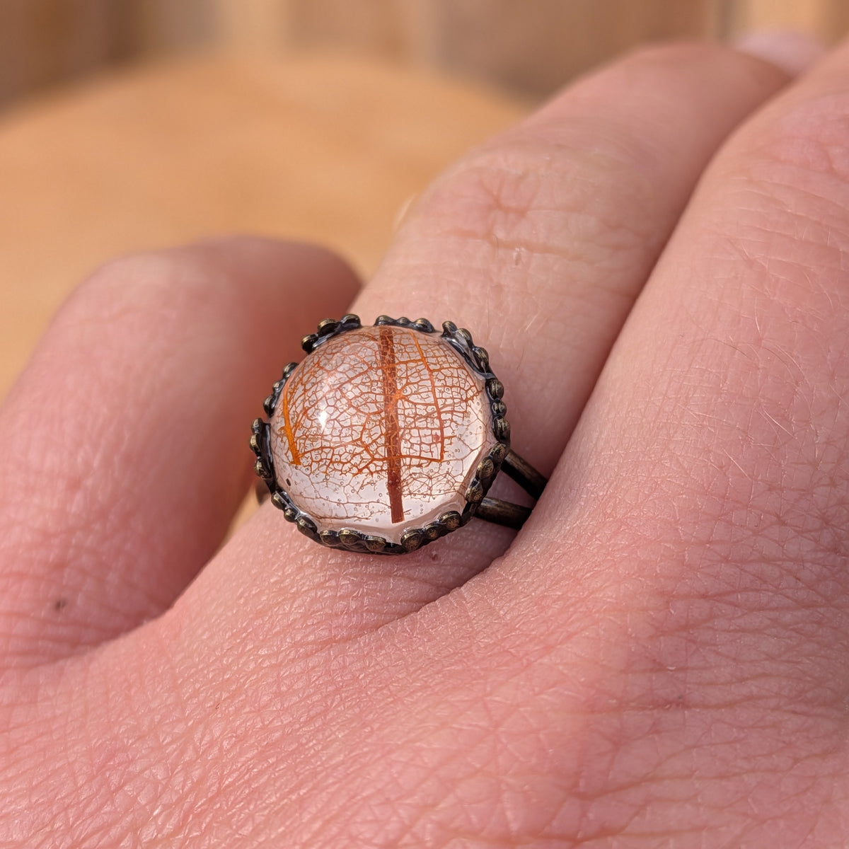 Close-up of an Autumn Glow resin ring worn on a finger, showing fine orange leaf veins beneath the domed surface, EL-Creations