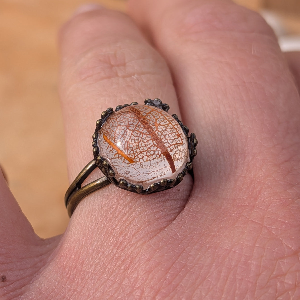 Angled view of an orange and beige botanical resin ring worn on a finger, highlighting its raised cabochon and brown split band, EL-Creations