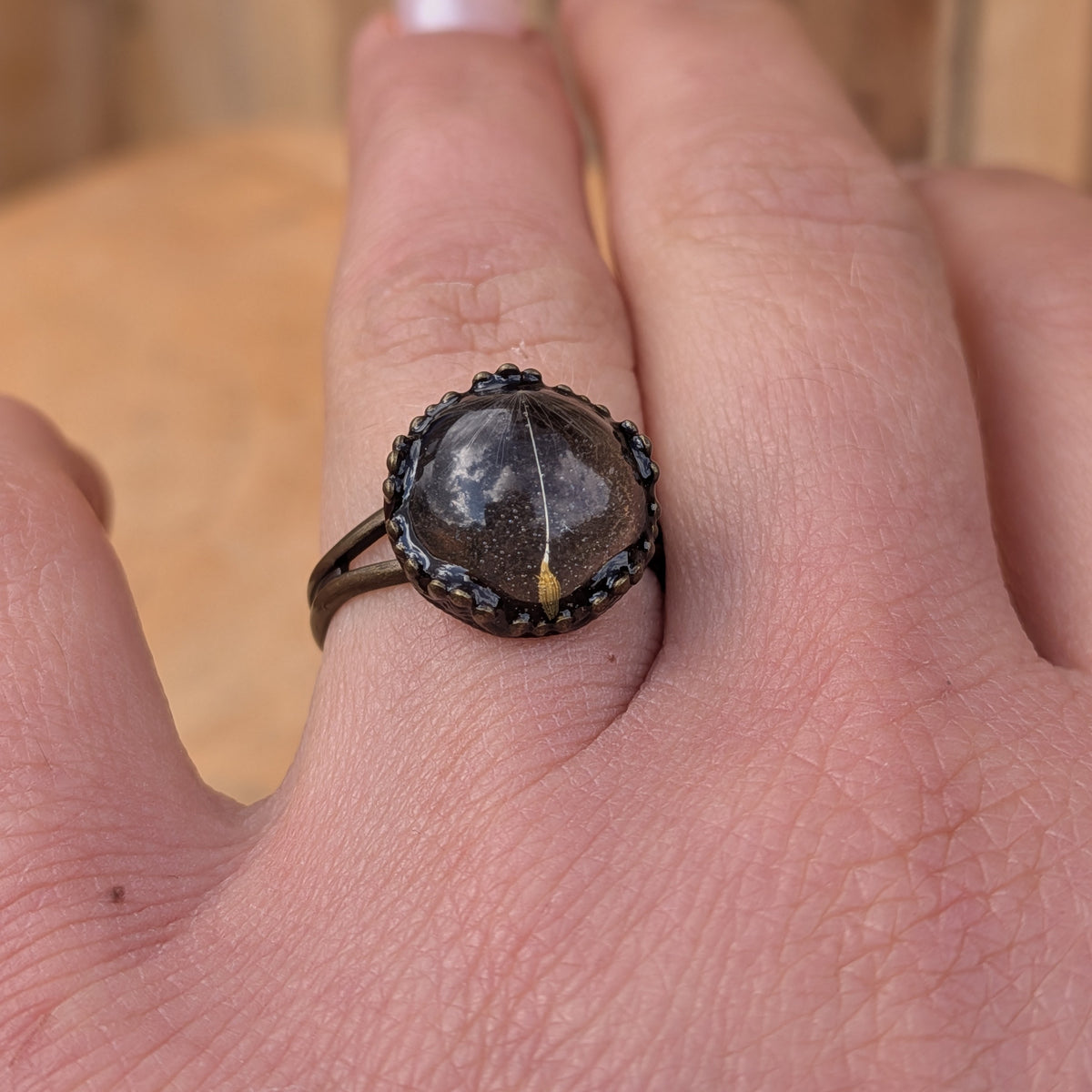 Front view of an adjustable dandelion seed resin ring worn on a finger, with its rustic brown split band visible beside the round centre, EL-Creations