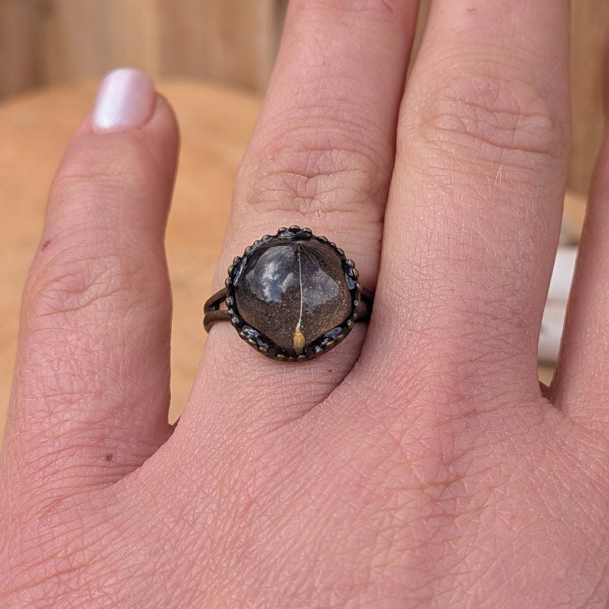 One Little Wish botanical resin ring worn on an outstretched hand, showing a single dandelion seed in a decorative brown setting, EL-Creations