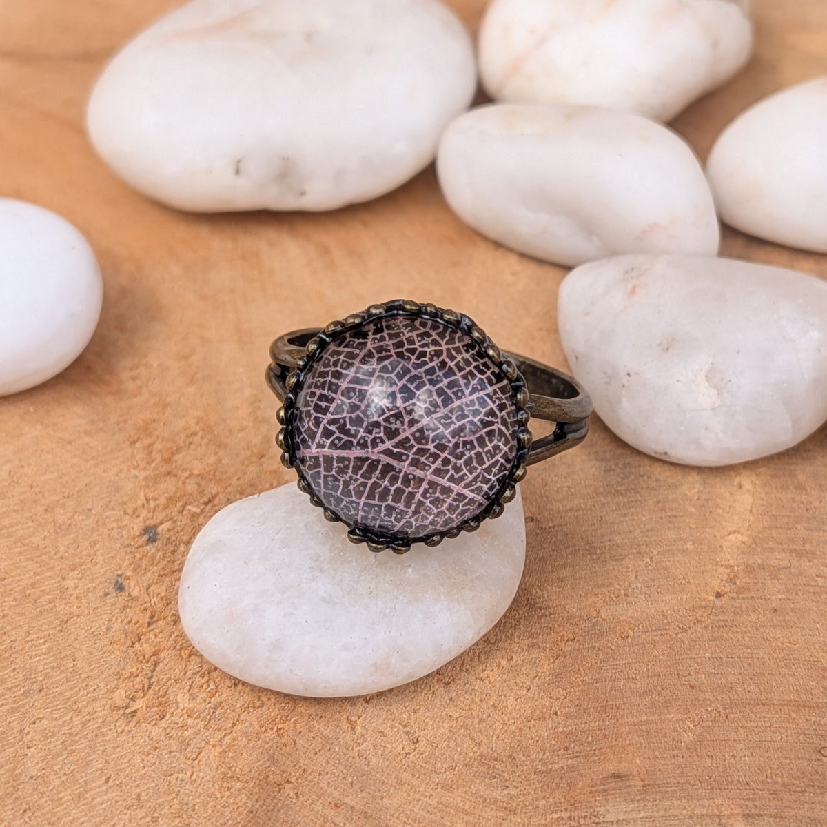 Close-up of a pale pink skeleton leaf resin ring among white pebbles, framed by a decorative rustic brown border, EL-Creations