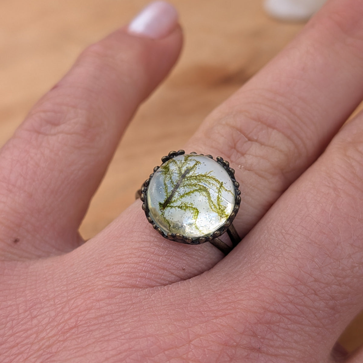 Angled close-up of a Moss Haven resin ring worn on a finger, showing fine green moss branches beneath the glossy surface, EL-Creations