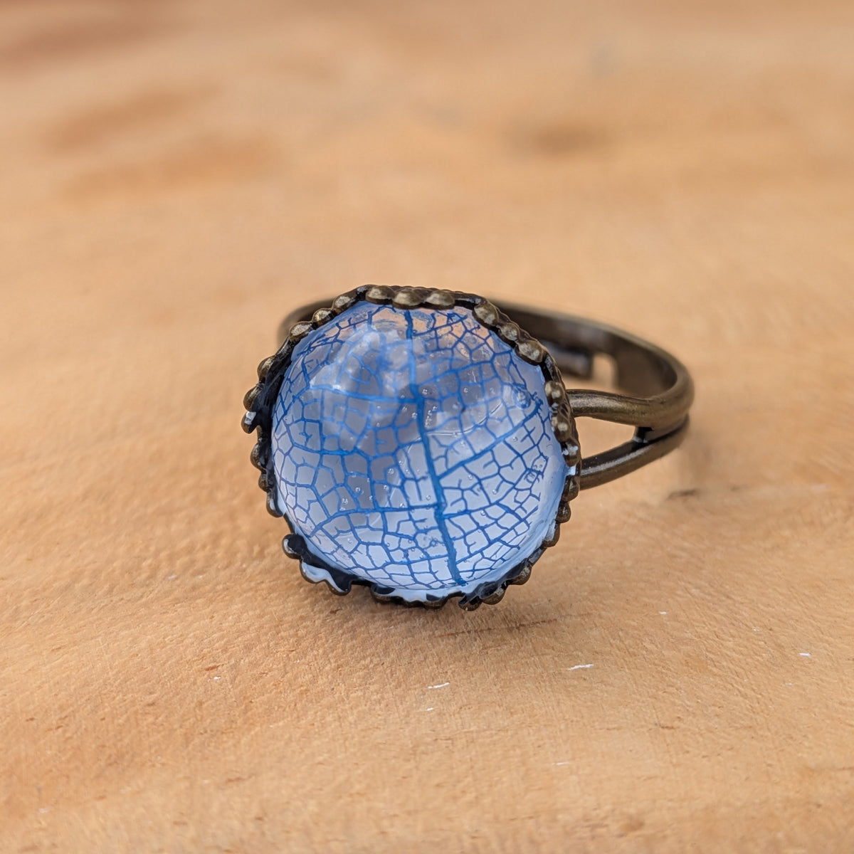 Macro view of rich blue skeleton leaf veins beneath a glossy resin dome, framed by a rustic brown ring setting on wood, EL-Creations