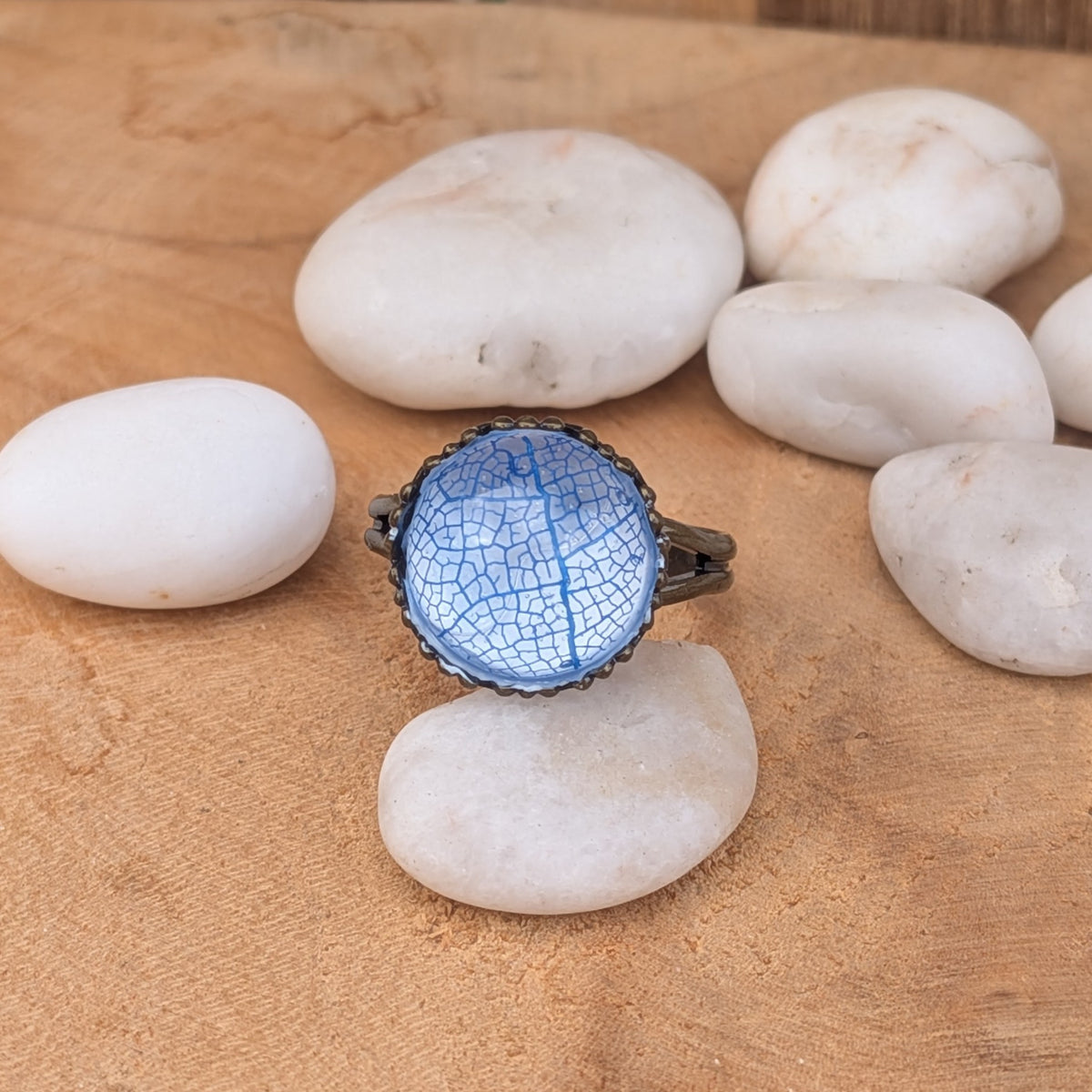 Front view of an Azure Lace blue leaf resin ring balanced on a white pebble among smooth stones, EL-Creations