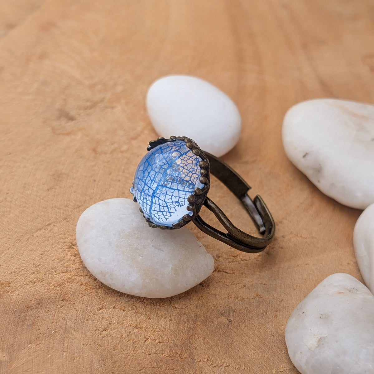 Side view of a blue botanical resin ring on a white stone, showing its open adjustable brown band and raised cabochon, EL-Creations