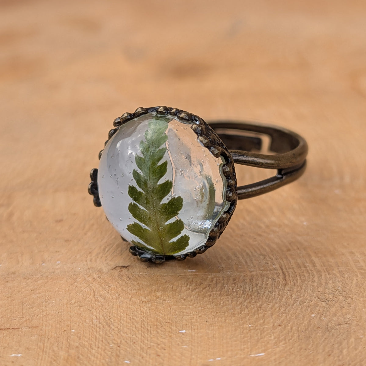 Angled macro view of a real green fern resin ring on wood, showing its raised cabochon and rustic brown split band, EL-Creations