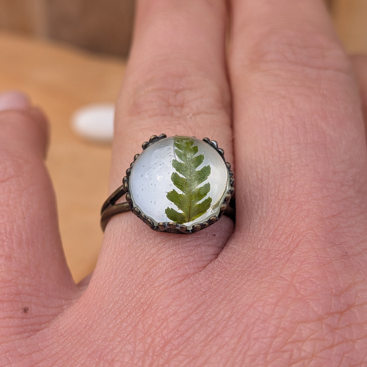 Front close-up of a Fern Whisper resin ring worn on a finger, showing a real green fern frond in a decorative rustic brown setting, EL-Creations