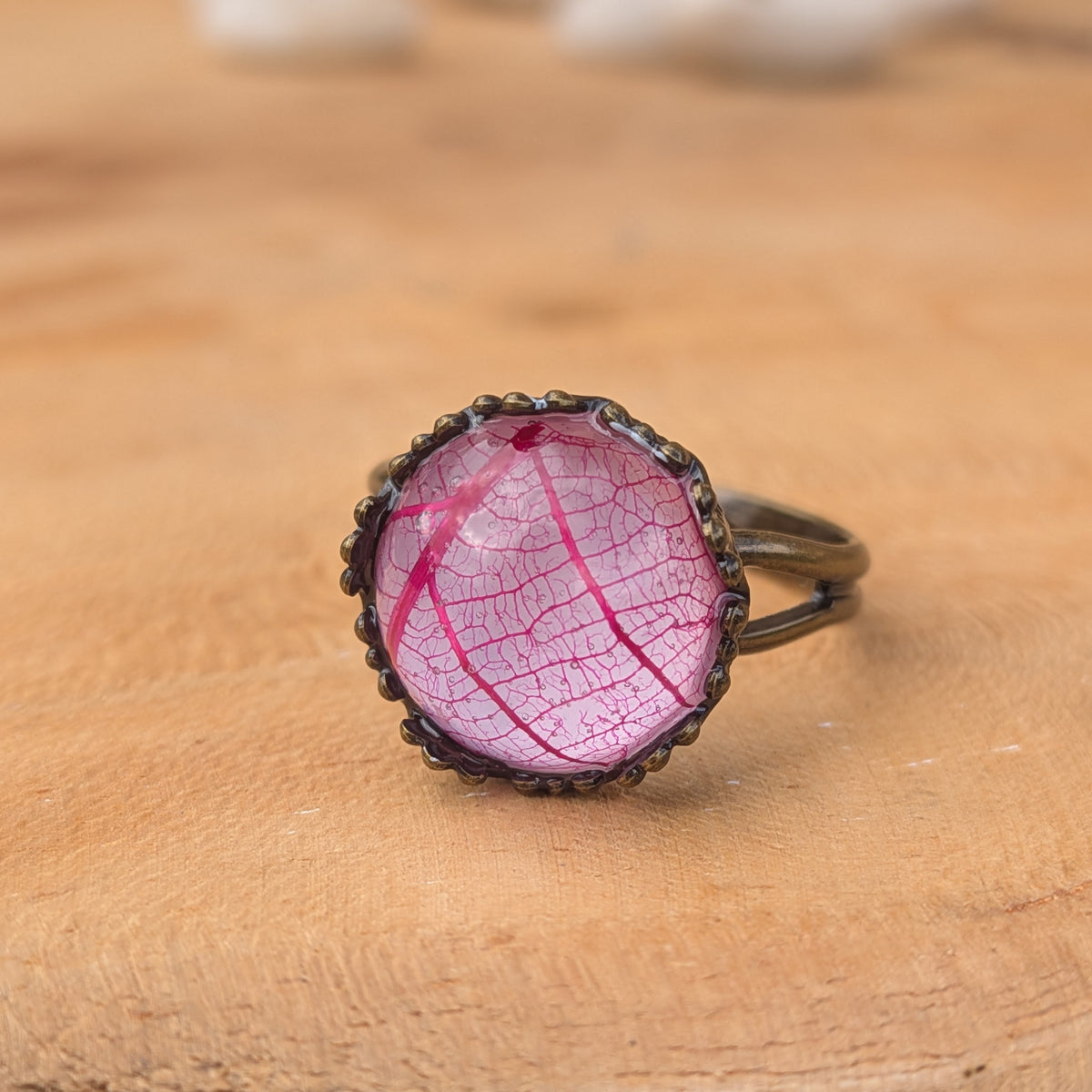 Macro view of a pink skeleton leaf preserved in resin, framed by a decorative brown ring setting on wood, EL-Creations