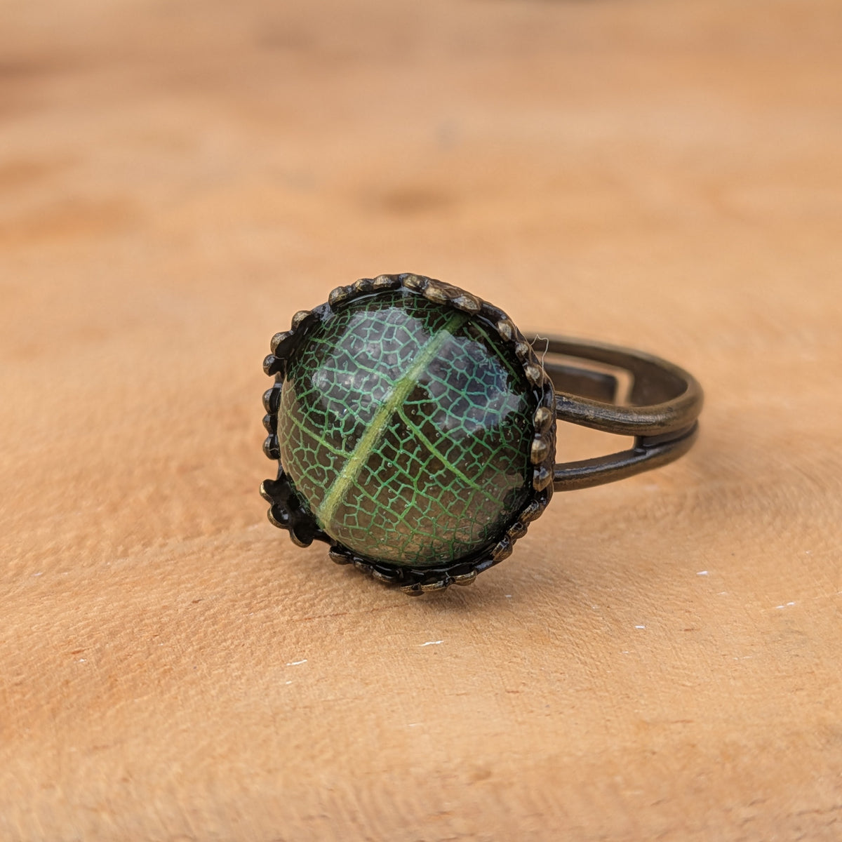 Green botanical resin ring resting directly on wood, showing its lace-like leaf pattern and adjustable brown split band, EL-Creations
