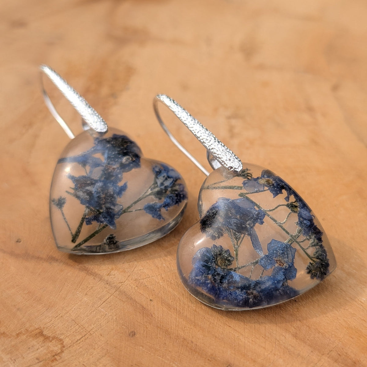 Forget-me-not heart earrings held against a blue sky showing floating flowers by EL-Creations